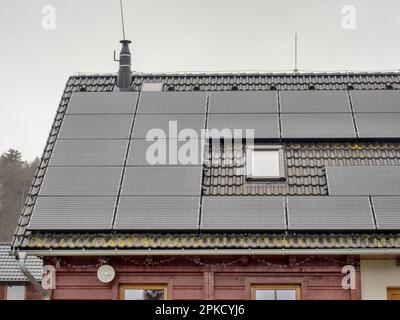 Sonnenkollektor auf schwarzem Dach mit kleinen Dachfenstern gegen bewölkten Himmel. Dunkler Regentag. Stockfoto