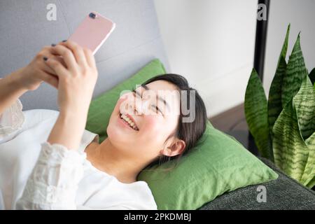 Frau lächelt, während sie ein Smartphone bedient, während sie auf dem Sofa liegt Stockfoto