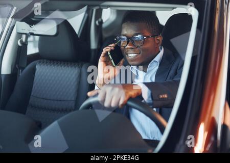 Mit dem Telefon. Ein junger afroamerikanischer Geschäftsmann im schwarzen Anzug sitzt im Auto. Stockfoto