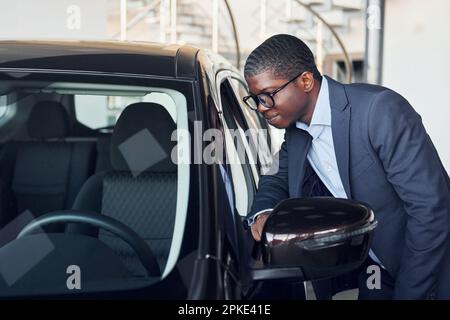 Ich öffne die Tür. Der junge afroamerikanische Geschäftsmann im schwarzen Anzug ist der autosalon Stockfoto