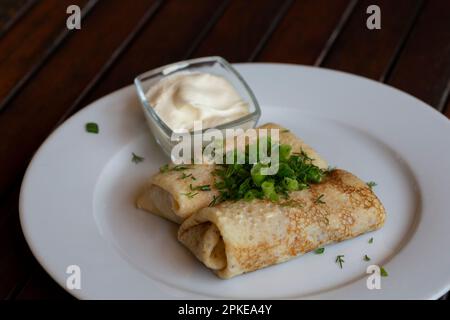 Zwei Hälften Pfannkuchenrolle mit Himbeerfüllung, saure Sahne auf weißem Teller, Teelöffel in Schüssel mit saurer Sahne, Gabel auf Holztisch, selektiver Fokus Stockfoto