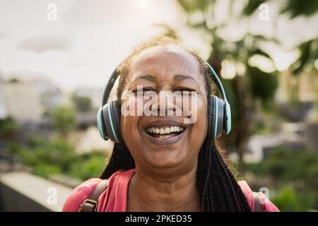 Glückliche afrikanische Seniorin, die Musik mit kabellosen Kopfhörern hört und gleichzeitig in die Kamera lächelt Stockfoto