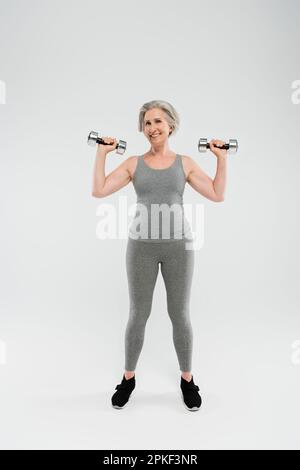 Volle Länge einer glücklichen Seniorin in enger Sportbekleidung mit Kurzhanteln auf grauem Stockbild Stockfoto