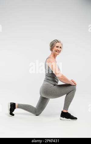 Die ganze Länge einer glücklichen Seniorin in Sportbekleidung, die Ausfallschritte macht und lächelt auf grauem Stockbild Stockfoto