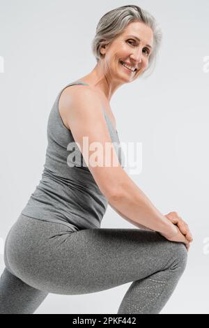 Glückliche Seniorin in Sportbekleidung, die Ausfallschritte macht und lächelt, isoliert auf grauem Stockbild Stockfoto