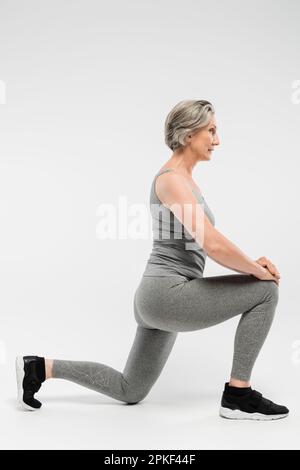 Die ganze Länge einer Seniorin in Sportbekleidung, die Ausfallschritte auf grauem Stockbild macht Stockfoto