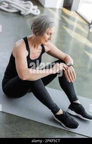 Lange, fit Seniorin in schwarzen Leggings und Tanktop, die den Fitness-Tracker überprüft, während sie auf der Fitnessmatte sitzt, Stockbild Stockfoto