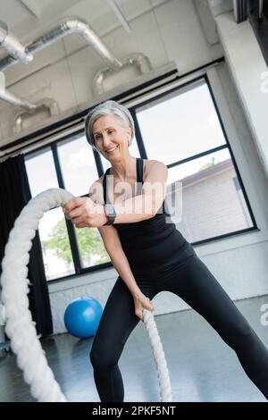 Fröhliche ältere Sportlerin mit grauen Haaren, die mit Kampfseilen im Fitnessstudio trainiert, Stockbild Stockfoto