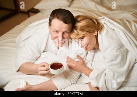 Ein Paar mittleren Alters in Bademänteln, das sich im Bett entspannt Stockfoto