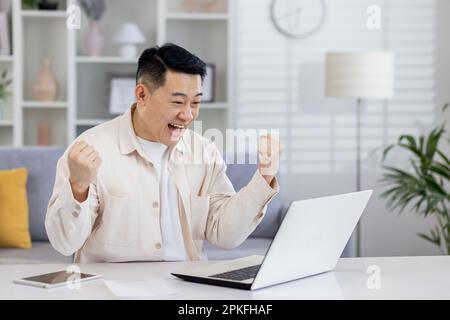 Asiaten, die von zu Hause aus mit einem Notebook arbeiten, erhielten eine Benachrichtigung über gute Erfolge, ein mit der Arbeit zufriedener Freiberufler freut sich und feiert Sieg und Triumph. Stockfoto
