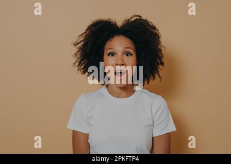 Foto einer verblüfften, dunkelhäutigen Frau, die mit offenem Mund in die Kamera schaut, einer schockierten afrikanischen Frau in einem weißen T-Shirt mit überraschtem Gesichtsausdruck Stockfoto