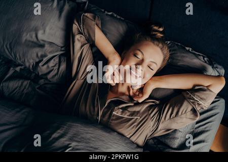Guten Morgen. Blick von oben auf die junge, wunderschöne, entspannte, brünette Frau mit geschlossenen Augen in Satin-Pyjamas, die in ihrem Bett vollständig ausgeruht aufwacht, glücklich wo Stockfoto