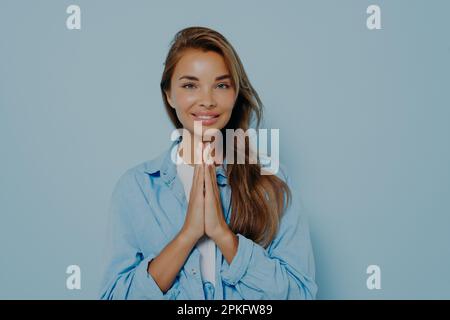 Fröhlich fröhlich angenehm aussehende Frau plädiert für etwas und hält die Handflächen zusammengedrückt, lächelt fröhlich, isoliert auf blauem Hintergrund. Schön Stockfoto