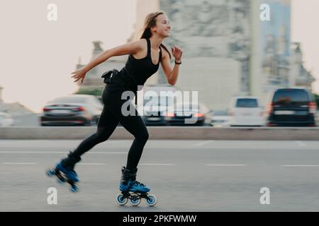 Junge dunkelhaarige schlanke gesunde Frau hat die Aufnahmezeit genießt Rollschuhlaufen auf Geschwindigkeit schnell hat fröhlichen Ausdruck trägt schwarzes T-Shirt und Leggin Stockfoto