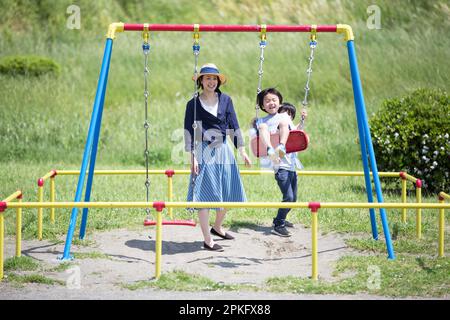 Der kleine Bruder auf der Schaukel mit dem älteren Bruder und der Mutter, die ihn schubsen Stockfoto