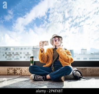 Speicherplatz kopieren. Wunderschönes Teenager-Mädchen, Internet-Influencer sitzt auf der Terrasse auf dem Balkon, benutzt Smartphone, macht ein Selfie und Unterhaltung. Gelb Stockfoto