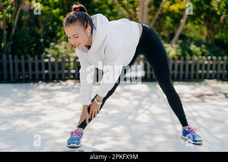Motivierte junge Erwachsene Frau geht zum Sport und erreicht regelmäßig Hand zu Fuß. Neigt sich nach unten mit den Füßen schulterbreit auseinander trägt Sportkleidung und trainiert gern Stockfoto