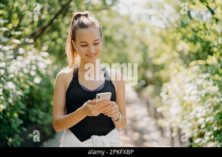 Sportliches Lifestyle- und Workout-Konzept. Junge, zufriedene sportliche Frau verwendet Smartphone, um online zu chatten und Musik zu hören, nachdem sie Kardio-Training ha Stockfoto