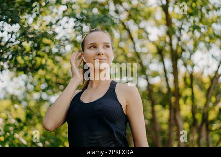 Die horizontale Aufnahme einer zufriedenen jungen Frau hält den Ohrstöpsel in der Hand Hört sich Lieblingsmusik an Looks Away hat einen befriedigenden Gesichtsausdruck in aktiver Kleidung gekleidet Stockfoto