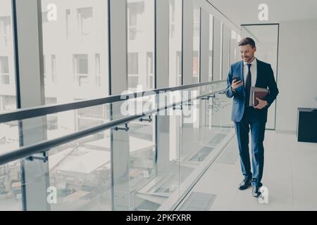 Lächelnder rothaariger Büroangestellter im Anzug, der sein Handy benutzt, E-Mails oder Nachrichten von einem Konto in einem sozialen Netzwerk abruft, Laptop und Notebook in der Hand hat, Klo Stockfoto