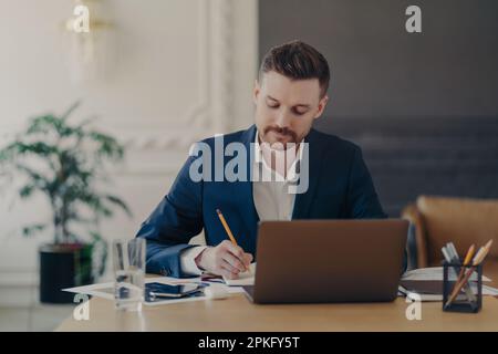 Seriöser, konzentrierter, geschulter Unternehmer prüft Finanzbuchhaltung Bericht schreibt einige Notizen in Notizblock sitzt in Teamarbeitsbereichen verwendet Laptop Com Stockfoto