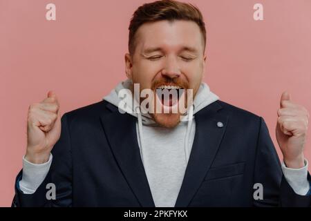 Studioaufnahme eines gutaussehenden, überfreulichen Rothaarigen, der Triumph feiert, überfreulicher, unrasierter Mann mit geschlossenen Augen, geballte Fäuste und Schreie Stockfoto