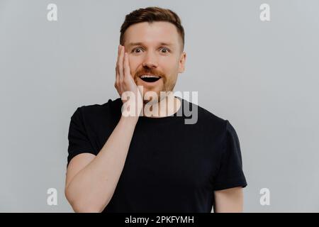 Ich kann es nicht glauben. Junger, gutaussehender Ingwermann in schwarzem T-Shirt, der den Kiefer aus der Aufregung herablässt, von etwas unglaublichem Whi schockiert zu werden Stockfoto