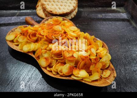 Frittierte, scharfe runde Scheiben neuer Kartoffeln auf traditionellem Holzteller auf schwarzem, öligen Kochfeld und knusprigem, flachgebackenem Brot auf der Lebensmittelmesse in Bukarest Stockfoto