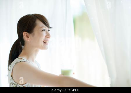 Eine Frau hält einen Smoothie und öffnet die Vorhänge Stockfoto