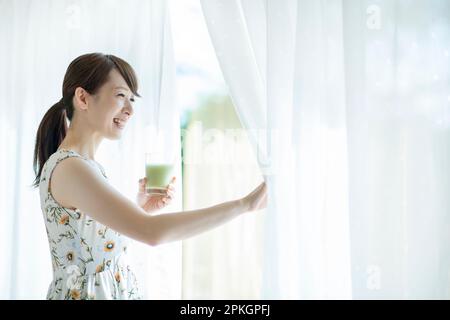 Eine Frau hält einen Smoothie und öffnet die Vorhänge Stockfoto