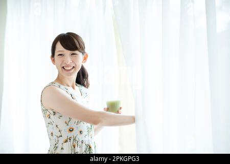 Eine Frau hält einen Smoothie und öffnet die Vorhänge Stockfoto