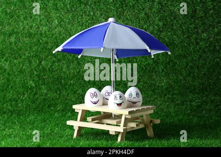 Eier mit fröhlichen Gesichtern, kleiner Picknicktisch und Sonnenschirm auf grünem Gras Stockfoto