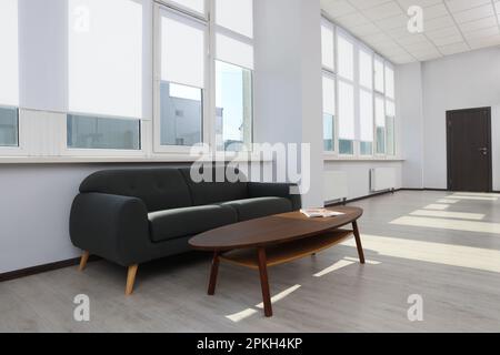 Komfortables Sofa und Tisch in der Nähe großer Fenster mit weißen Rollläden im Innenbereich Stockfoto