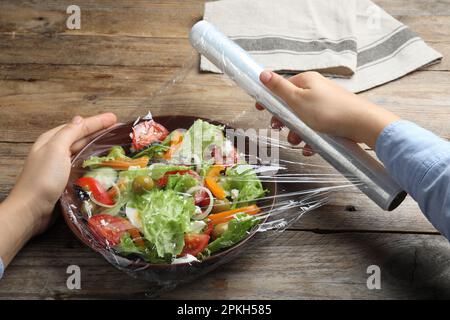 Frau, die Plastikfolie über eine Schüssel mit frischem Salat am Holztisch legt, Nahaufnahme Stockfoto