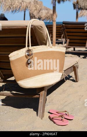 Strohtasche mit Sonnenbrille auf einer Holzliege in der Nähe des Meeres. Strandaccessoires Stockfoto