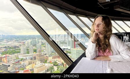 Ein junges Mädchen im Teenageralter sitzt am Fenster eines sich drehenden Restaurants und ist traurig, dass sie ihre Hand neben ihrem Gesicht traurig ist und sie aus dem Fenster auf das Visights Top of Vancouver Revolving Restaurant blickt. 4K Stockfoto