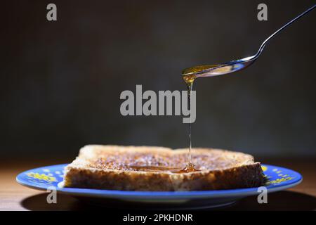 Eine Seitenansicht eines geneigten Löffels, der frischen Honig auf eine getoastete Scheibe Brot tropft, auf einem Teller, in sanfter, stimmungsvoller Beleuchtung mit Kopierbereich auf der linken Seite Stockfoto