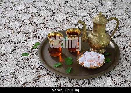 Eine klassische, feierliche, kunstvoll verzierte türkische Teekanne, zwei Gläser und traditionelle türkische Köstlichkeiten auf einem Tablett und weiße Tischdecke aus Spitze in sanfter Beleuchtung Stockfoto