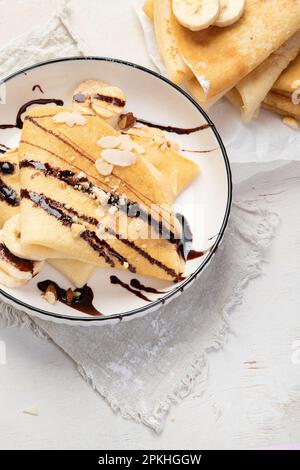 Crepe mit Banane, gehackten Mandeln und Schokoladensoße auf weißem Teller, Blick vom Tisch auf weißem Hintergrund. Köstliche crepes nach französischer Art. Stockfoto