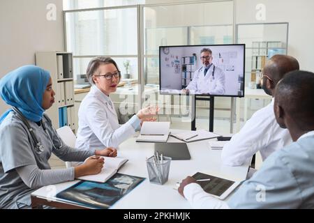 Eine Gruppe von Ärzten führt ein Videogespräch mit einem Kollegen am Monitor. Sie sitzen am Tisch und besprechen Arbeitsmomente während des Meetings im Büro Stockfoto