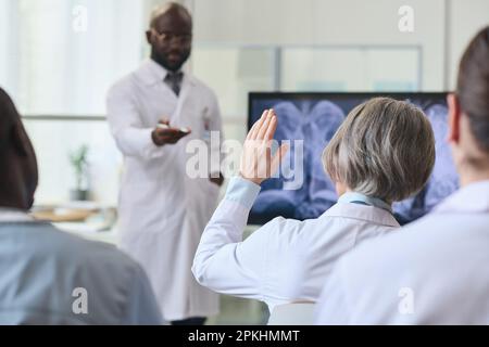 Rückansicht einer Ärztin im weißen Kittel, die ihre Hand hebt und Fragen während der medizinischen Konferenz mit ihren Kollegen stellt Stockfoto