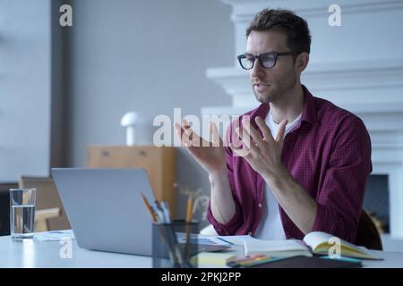 Junger fokussierter deutscher Unternehmer in Brillen und Freizeitkleidung, der an einem virtuellen Online-Meeting mit Kollegen teilnimmt und Videoanrufe auf Schoß führt Stockfoto