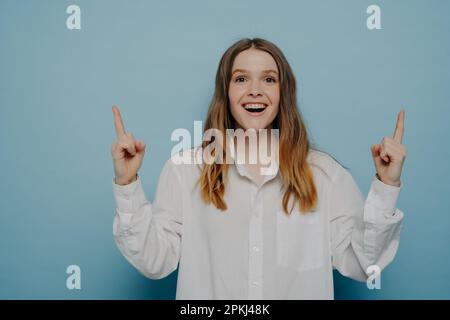 Erstaunte junge Frau, die mit den Fingern nach oben zeigt und Aufregung ausdrückt, während sie etwas erstaunliches sieht, isoliert an der blauen Wand, was eine großartige Oper bietet Stockfoto