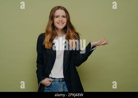 Lächelnde junge Frau mit gebeugter Hand, die Verwirrung und Unsicherheit demonstriert und eine dunkle formelle Jacke, ein weißes Oberteil und Jeans trägt, die isoliert über Gre stehen Stockfoto