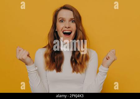 Junge, emotional betäubte Schülerin, die einen beeindruckenden Gesichtsausdruck macht und die Fäuste zusammendrückt, während sie im Studio posiert, isoliert auf gelbem Hintergrund, weiblich W. Stockfoto