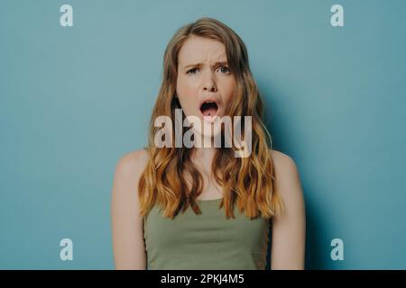 Es ist schrecklich. Junge unzufriedene Frau mit leicht welligem Haar, die auf die Kamera schaut, mit unzufriedenem, schockiertem Gesichtsausdruck und offenem Mund, unglückliche Frauen Stockfoto