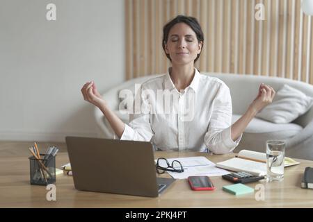 Ruhige italienische Frau sitzt in entspannter Yoga-Position am Schreibtisch vor dem Laptop mit geschlossenen Augen, während sie ihre Finger in Mudra faltet, um sich besser zu konzentrieren Stockfoto