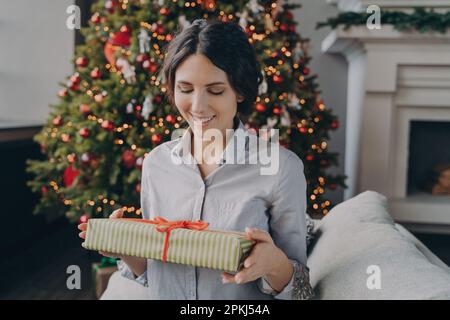 Lächelnde europäische Frau, die auf dem Sofa sitzt, in einem für Weihnachtsfeiertage dekorierten Zimmer mit eingepacktem Weihnachtsgeschenk, glückliche hispanische Frau, die auf Geschenk bo schaut Stockfoto