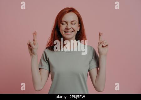 Studiofoto einer hoffnungsvollen rothaarigen Studentin mit geschlossenen Augen in lässiger Kleidung mit abergläubischen Gesichtsausdrücken, die nach Glibber fragt Stockfoto