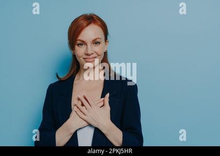 Studioaufnahme einer glücklichen lächelnden Frau mit rotem Haar, die Hände am Herzen hält und dankbar, dankbar ist, eine Geste der Dankbarkeit zeigt, während sie steht Stockfoto